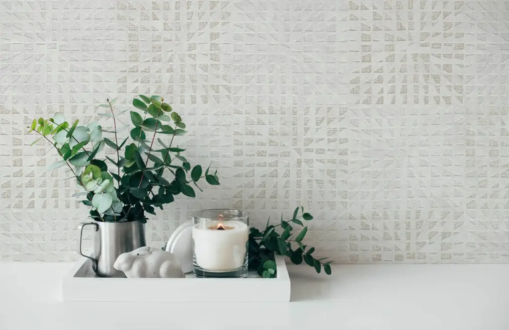 A close-up interior design shot showing a potted green plant, a candle, and decorative items on a white surface against a patterned wall.