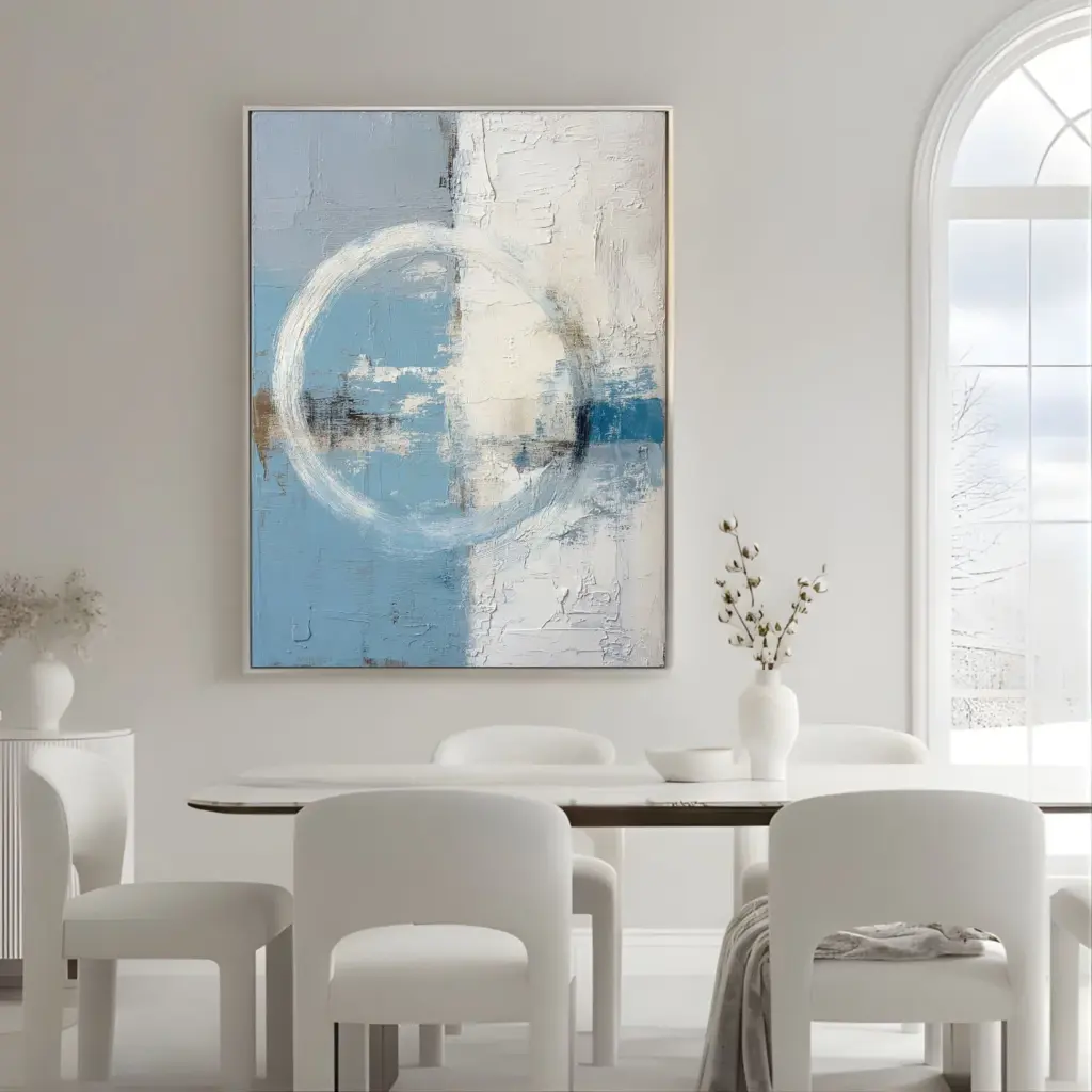 Abstract blue and white circular painting above dining table – A minimalist dining room with a white table and chairs, and a square abstract painting in soft blue and white circular patterns on the wall.